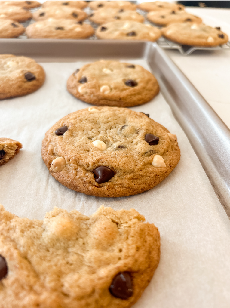 Golden brown cookies with dark and white chocolate chips on parchment paper, with one cookie bitten and more in the background.