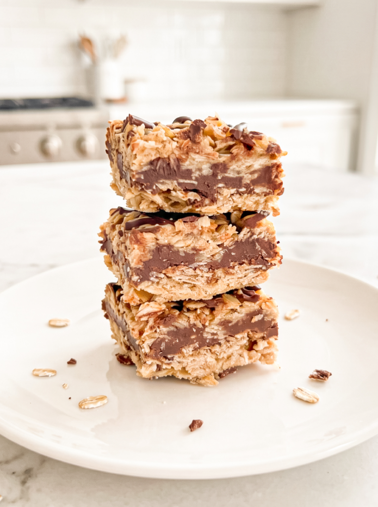 Three chunky chocolate oat bars with a thick fudge center are stacked on a white plate, with scattered oats and a blurry kitchen background.