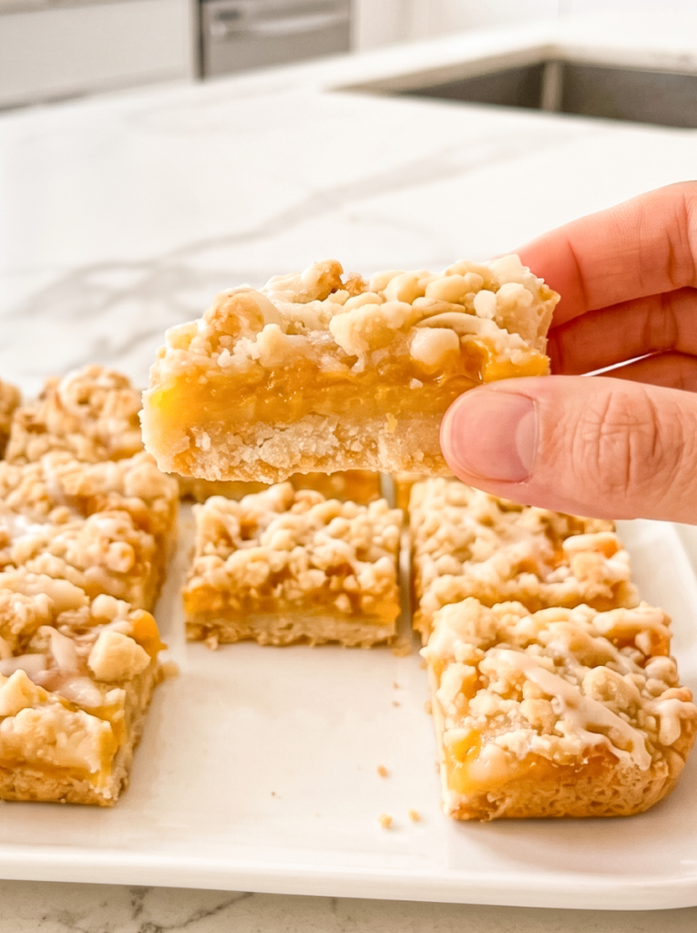 A hand holds a peach crumble bar with a thick peach filling, crumbly topping, and white glaze over more bars on a white platter.