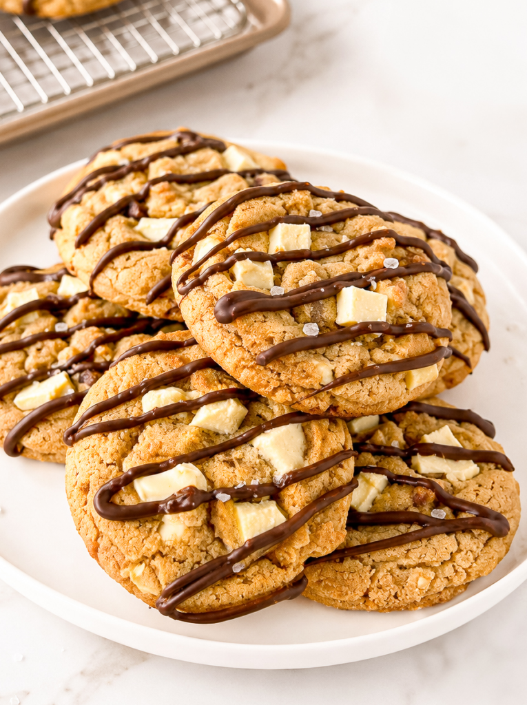 Peanut butter cookies with white chocolate chunks, drizzled with dark chocolate and sprinkled with sea salt, on a white plate.