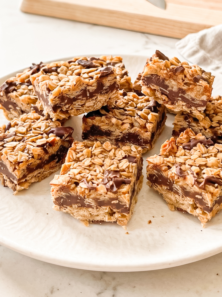 Several square no bake chunky choco oat bars with a thick chocolate layer and an oat topping are piled on a white textured plate.