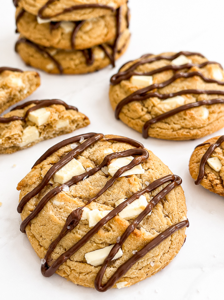 Peanut butter cookies with white chocolate chunks are drizzled with dark chocolate and sprinkled with sea salt on a white surface.