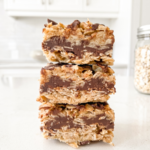 Three no-bake chunky chocolate oat bars with thick chocolate centers are stacked on a white countertop in a bright kitchen.