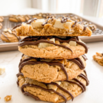 A stack of four peanut butter cookies with white chocolate chunks and chocolate drizzle, the top one split open, with more cookies on a cooling rack.