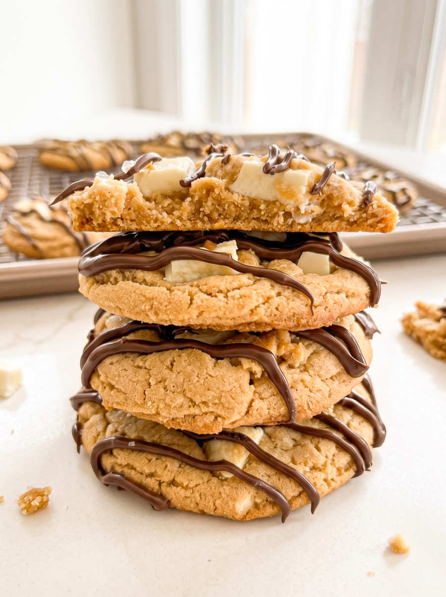 A stack of four peanut butter cookies with white chocolate chunks and chocolate drizzle, the top one split open, with more cookies on a cooling rack.
