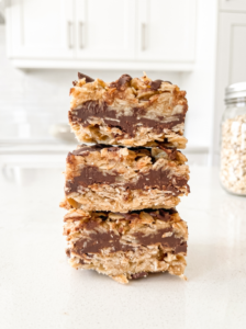 Three no-bake chunky chocolate oat bars with thick chocolate centers are stacked on a white countertop in a bright kitchen.