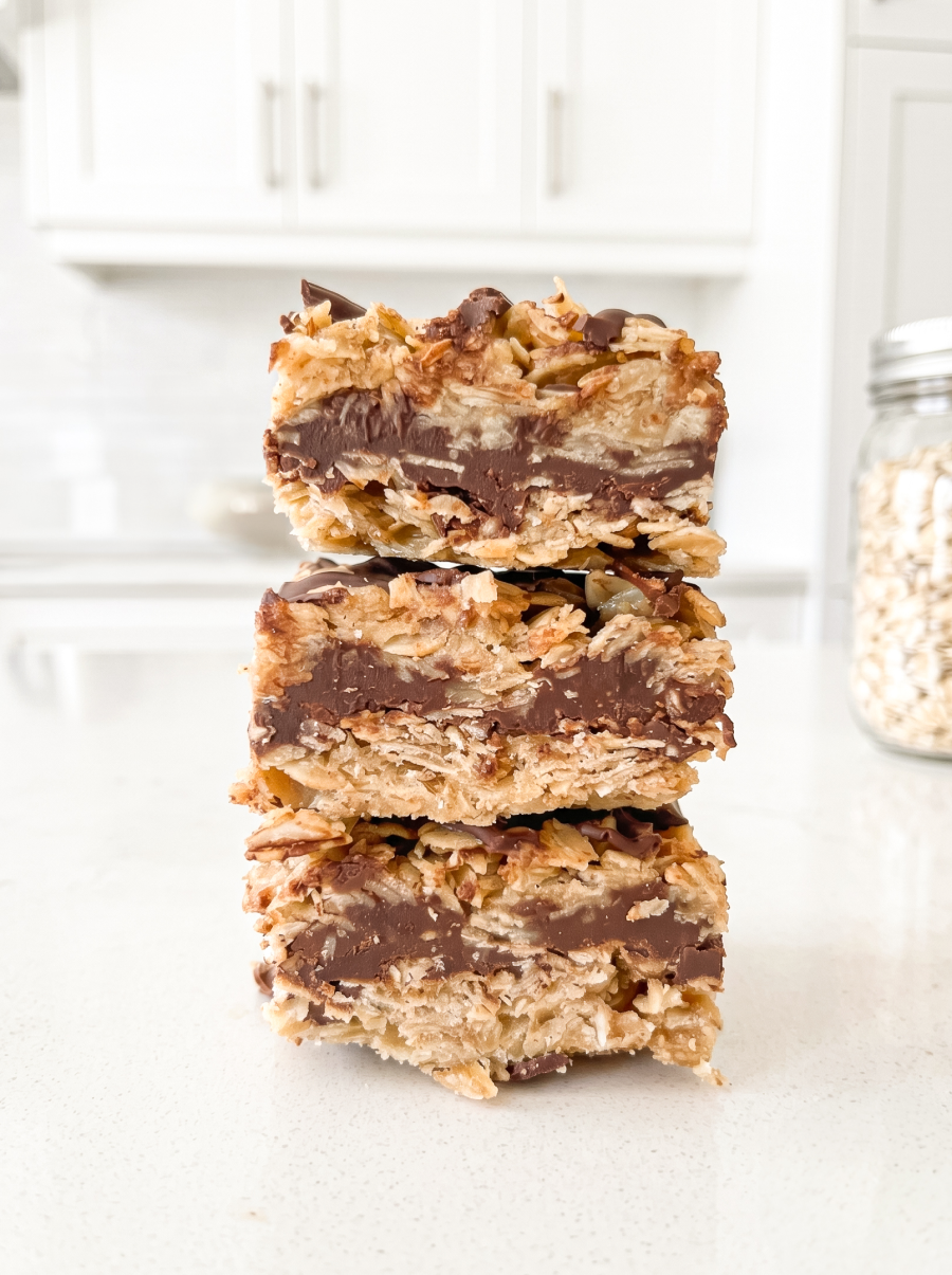 Three no-bake chunky chocolate oat bars with thick chocolate centers are stacked on a white countertop in a bright kitchen.