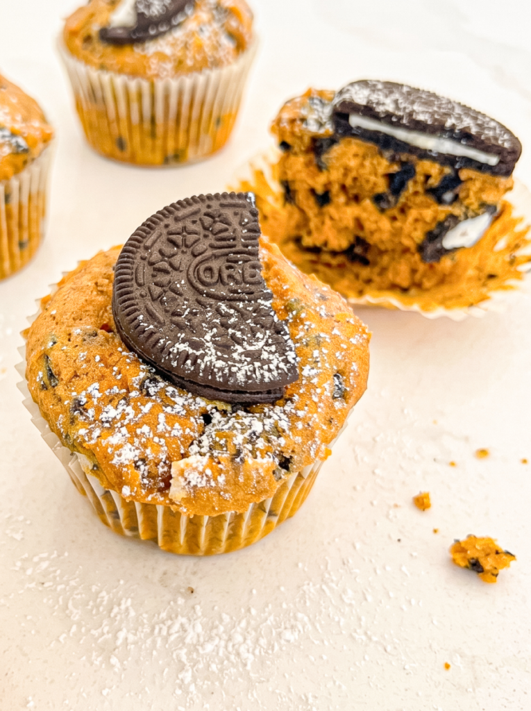 A pumpkin muffin in a white liner is dusted with icing sugar and topped with a half Oreo cookie, with a broken muffin behind it.