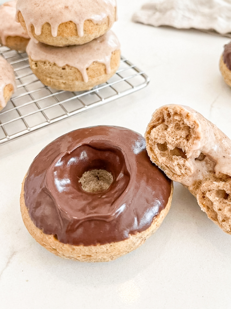 A chocolate-glazed donut next to a broken, light pink glazed donut, with more on a cooling rack behind.