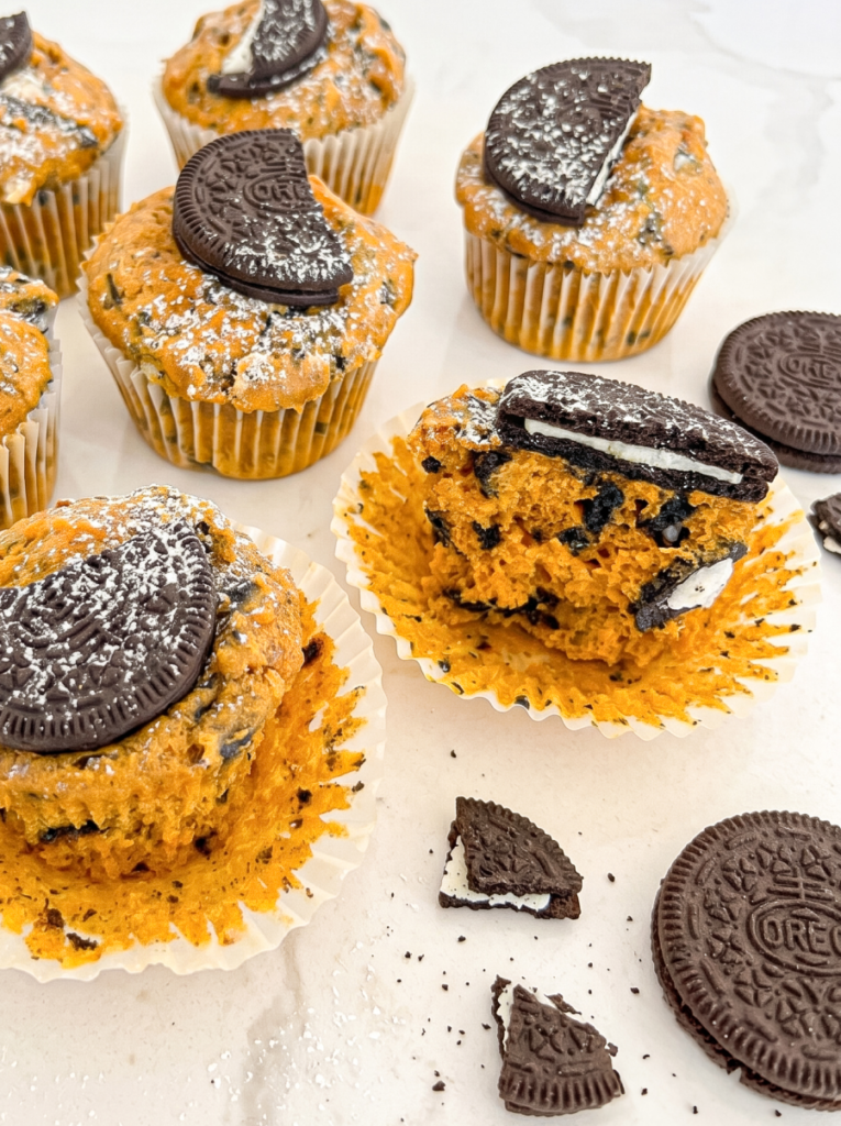 Six pumpkin muffins with crushed Oreos are topped with half an Oreo cookie and powdered sugar, one muffin is broken open.