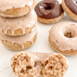 A stack of three vanilla chai donuts with dripping glaze sits next to a halved donut showing its fluffy texture, with more donuts in the background.