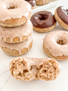 A stack of three vanilla chai donuts with dripping glaze sits next to a halved donut showing its fluffy texture, with more donuts in the background.