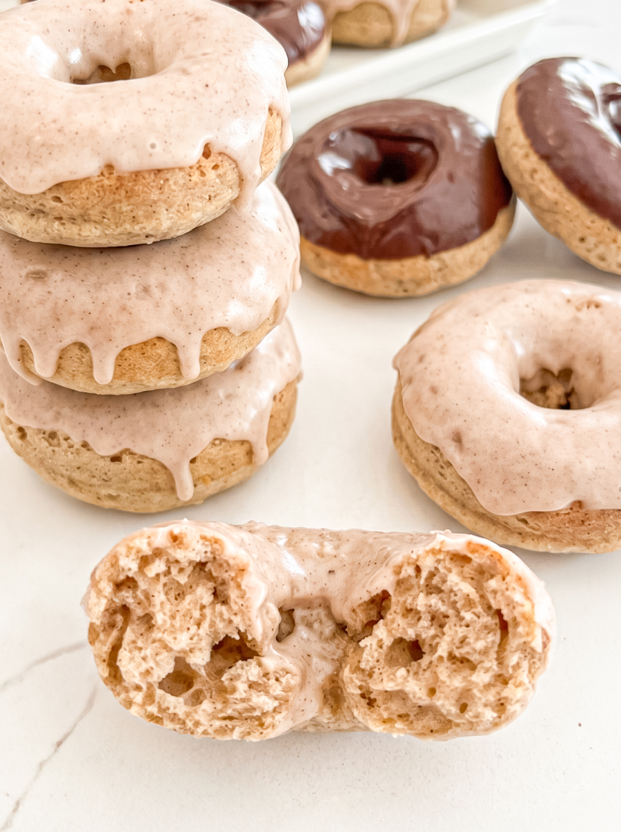 A stack of three vanilla chai donuts with dripping glaze sits next to a halved donut showing its fluffy texture, with more donuts in the background.
