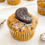 A pumpkin muffin with dark cookie pieces, powdered sugar, and a half Oreo cookie on top sits on a white surface.