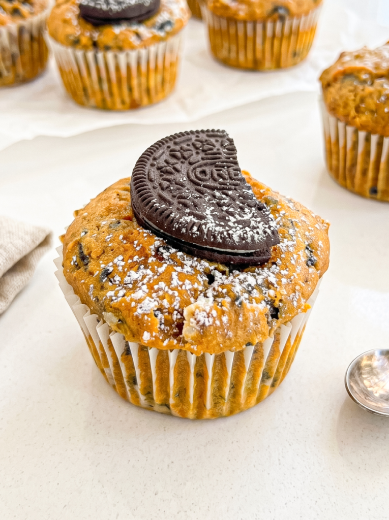 A pumpkin muffin with dark cookie pieces, powdered sugar, and a half Oreo cookie on top sits on a white surface.