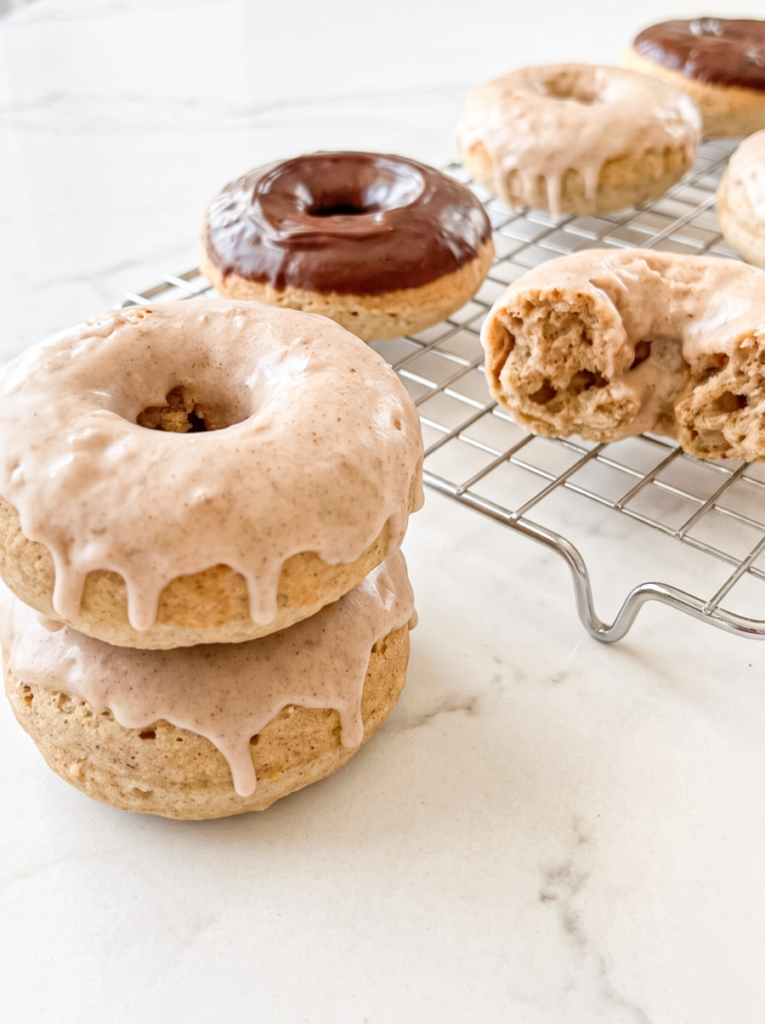 A stack of two glazed chai donuts sits beside a chocolate-frosted donut and a broken chai donut on a cooling rack over marble.