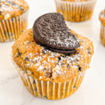 A close-up shows a pumpkin muffin with crushed Oreos, dusted with powdered sugar, and topped with a half Oreo cookie, with other muffins blurred in the background.