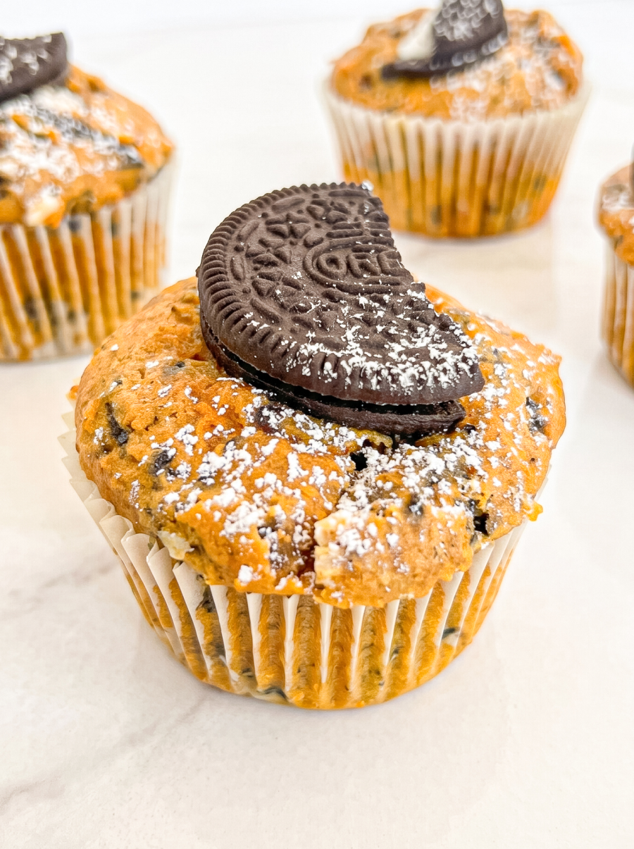 A close-up shows a pumpkin muffin with crushed Oreos, dusted with powdered sugar, and topped with a half Oreo cookie, with other muffins blurred in the background.