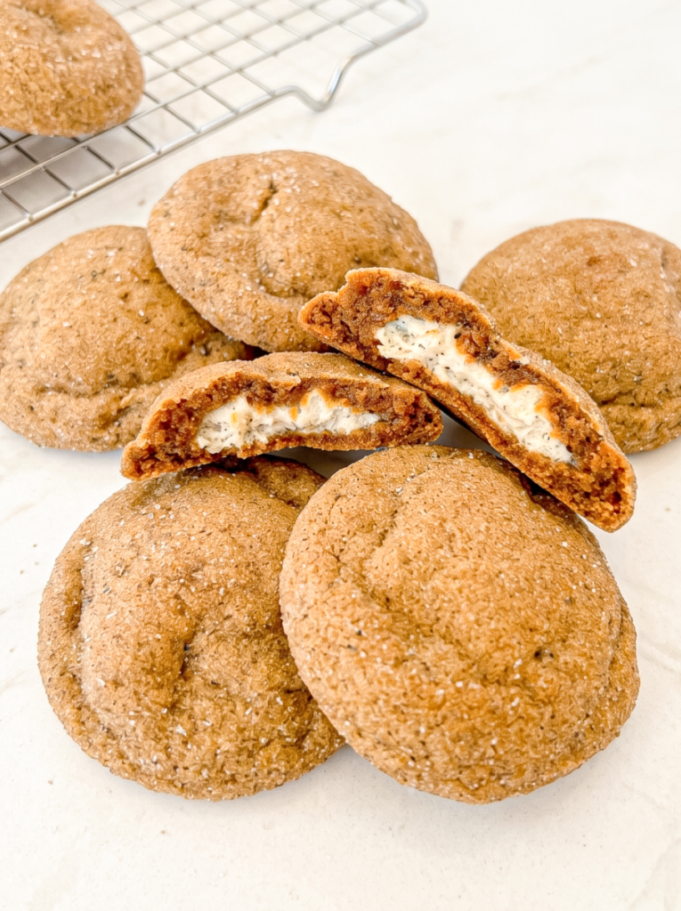 Several round, sugar-dusted cookies are on a white surface, with two broken to reveal a white, speckled cheesecake filling, and a cooling rack behind.