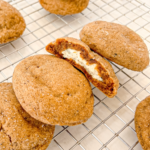 Several round, brown gingerbread chai cookies on a wire rack, one cut open to show a creamy cheesecake centre.
