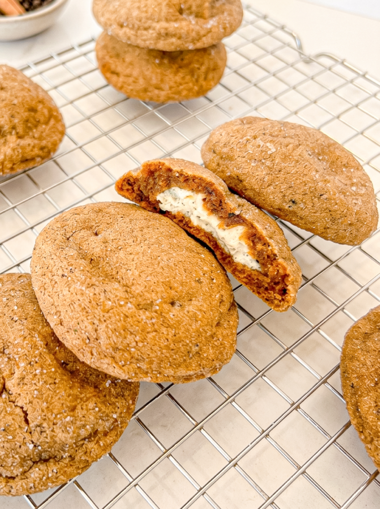 Several round, brown gingerbread chai cookies on a wire rack, one cut open to show a creamy cheesecake centre.