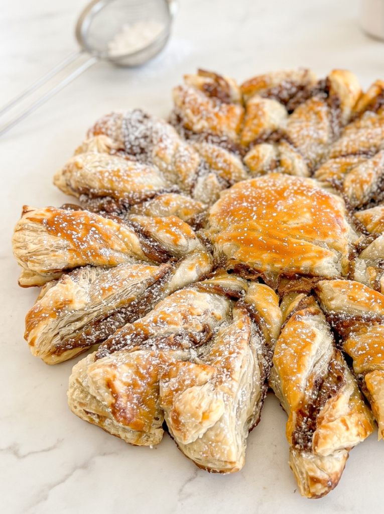 A golden-brown puff pastry snowflake filled with Nutella and Biscoff spread is dusted with icing sugar on a white surface.
