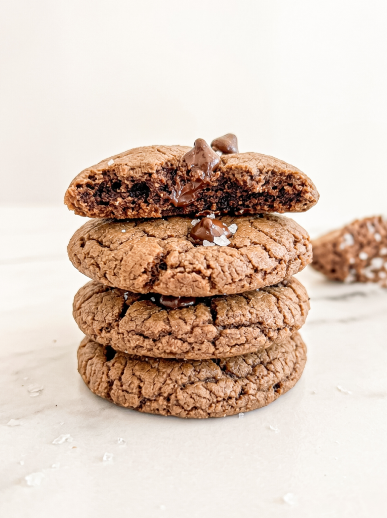 A stack of three brown cookies with chocolate chips and sea salt, the top cookie broken to show a melted chocolate centre.