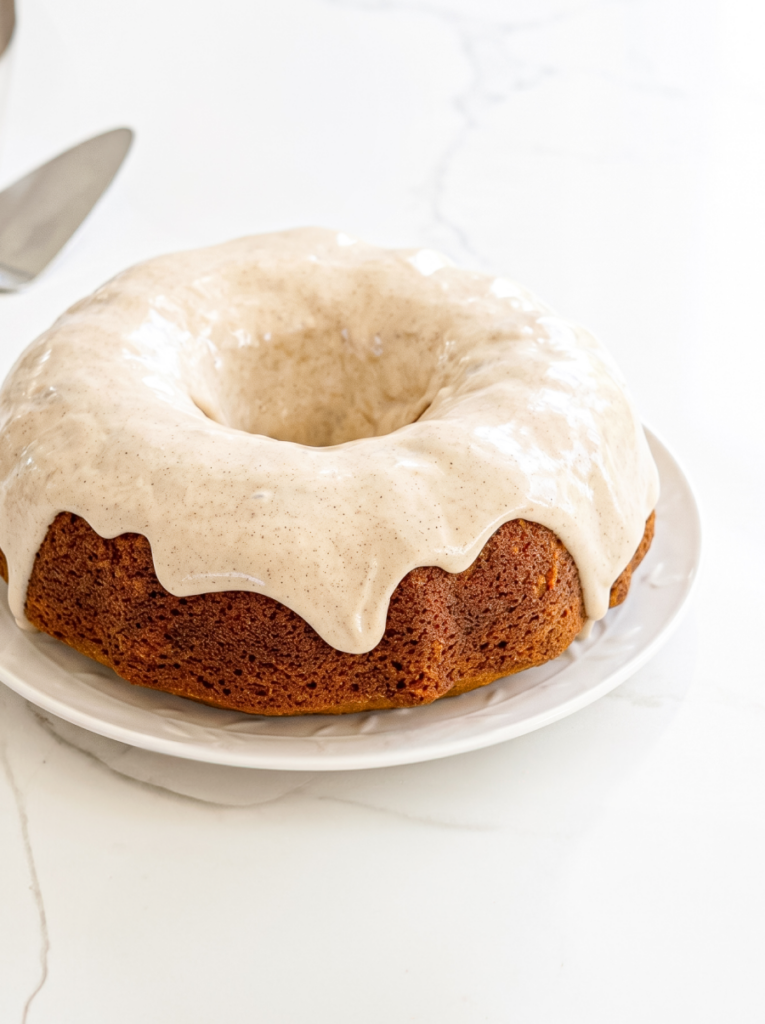 A golden brown bundt cake, topped with a light glaze that has visible spice flecks, sits on a white plate on a marble surface.