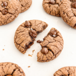 Chocolate cookies with crackled tops, chips, and sea salt are on a white surface; one cookie is broken in half, showing melted chocolate.