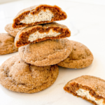 A stack of sugar-coated gingerbread cookies on a white surface, with two cookies broken open to show the white chai cheesecake filling.