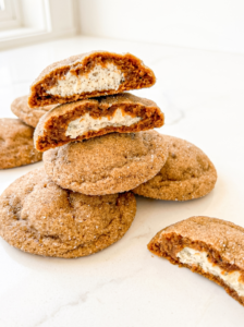 A stack of sugar-coated gingerbread cookies on a white surface, with two cookies broken open to show the white chai cheesecake filling.
