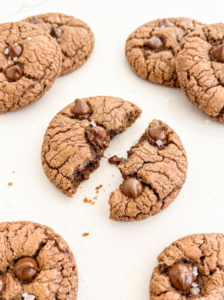 Chocolate cookies with crackled tops, chips, and sea salt are on a white surface; one cookie is broken in half, showing melted chocolate.