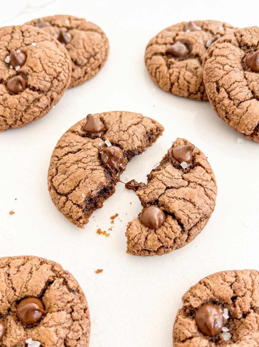 Chocolate cookies with crackled tops, chips, and sea salt are on a white surface; one cookie is broken in half, showing melted chocolate.