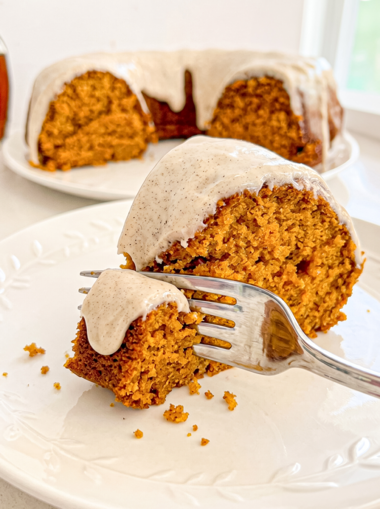 A slice of pumpkin maple bundt cake with a speckled glaze is on a white plate, with a fork taking a piece. The rest of the cake is in the background.