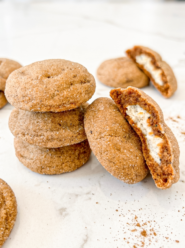 A stack of three round, light brown cookies with a sugar coating sits next to a split cookie revealing a white cream cheese filling.