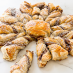 A golden-brown puff pastry in a snowflake shape, filled with dark brown spread and dusted with powdered sugar, sits on a white marble surface.