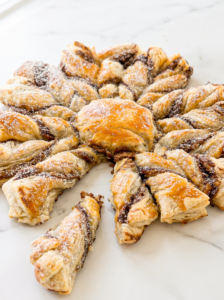 A golden-brown puff pastry in a snowflake shape, filled with dark brown spread and dusted with powdered sugar, sits on a white marble surface.