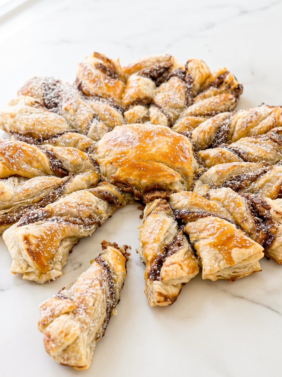 A golden-brown puff pastry in a snowflake shape, filled with dark brown spread and dusted with powdered sugar, sits on a white marble surface.