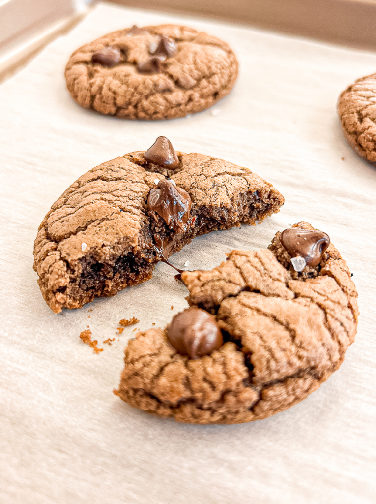 A brown crinkled cookie broken in half on parchment paper, showing a gooey chocolate centre with melted chocolate dripping out, surrounded by other cookies.