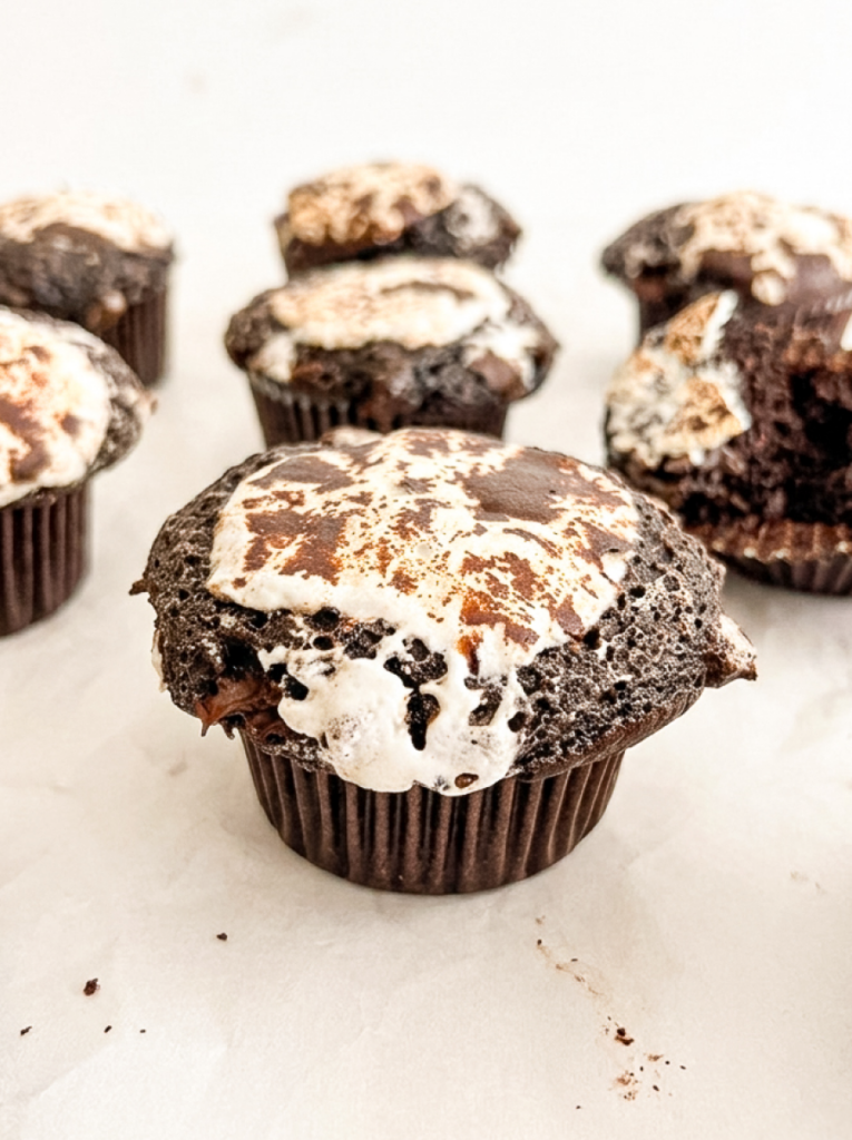 A dark chocolate muffin in a brown paper liner sits in the foreground, topped with browned, melted marshmallows.