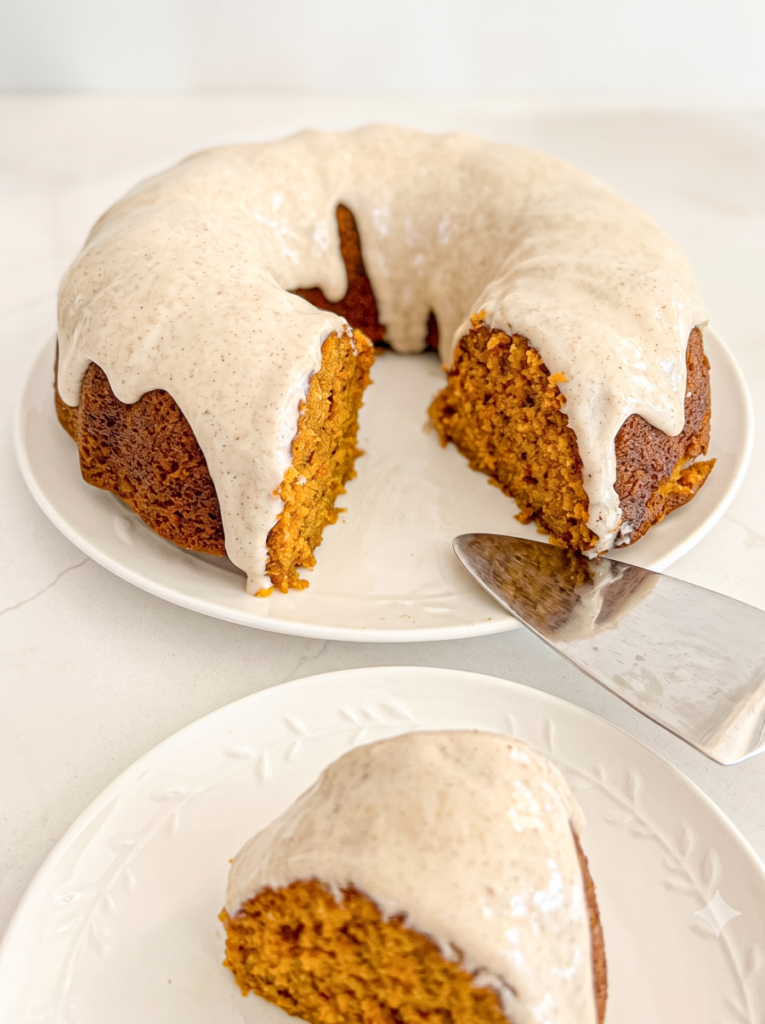 A pumpkin maple bundt cake with a light glaze drizzled over it, with one slice removed and placed on a smaller plate, beside the cake.