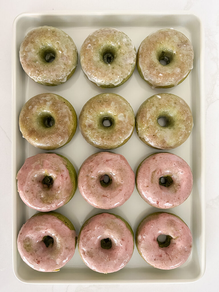 Dozen baked matcha donuts are arranged on a white tray, six with white glaze and six with pink glaze.