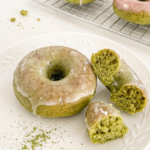 Glazed matcha donut, broken in half, on a white plate with matcha powder, beside more donuts on a wire rack.