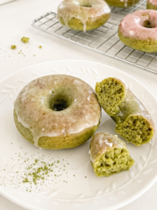 Glazed matcha donut, broken in half, on a white plate with matcha powder, beside more donuts on a wire rack.