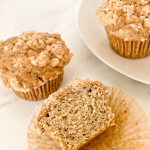 Two Earl Grey crumble muffins sit on a marble surface, with one cut in half showing its texture. Another whole muffin is on a white plate.