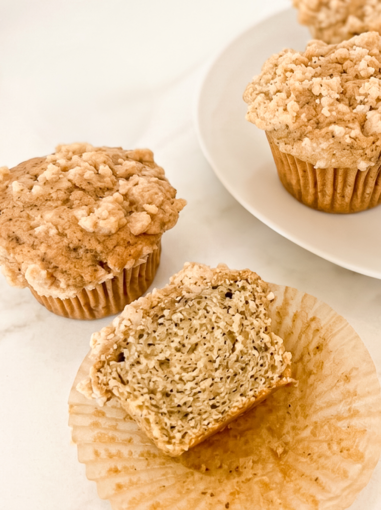 Two Earl Grey crumble muffins sit on a marble surface, with one cut in half showing its texture. Another whole muffin is on a white plate.