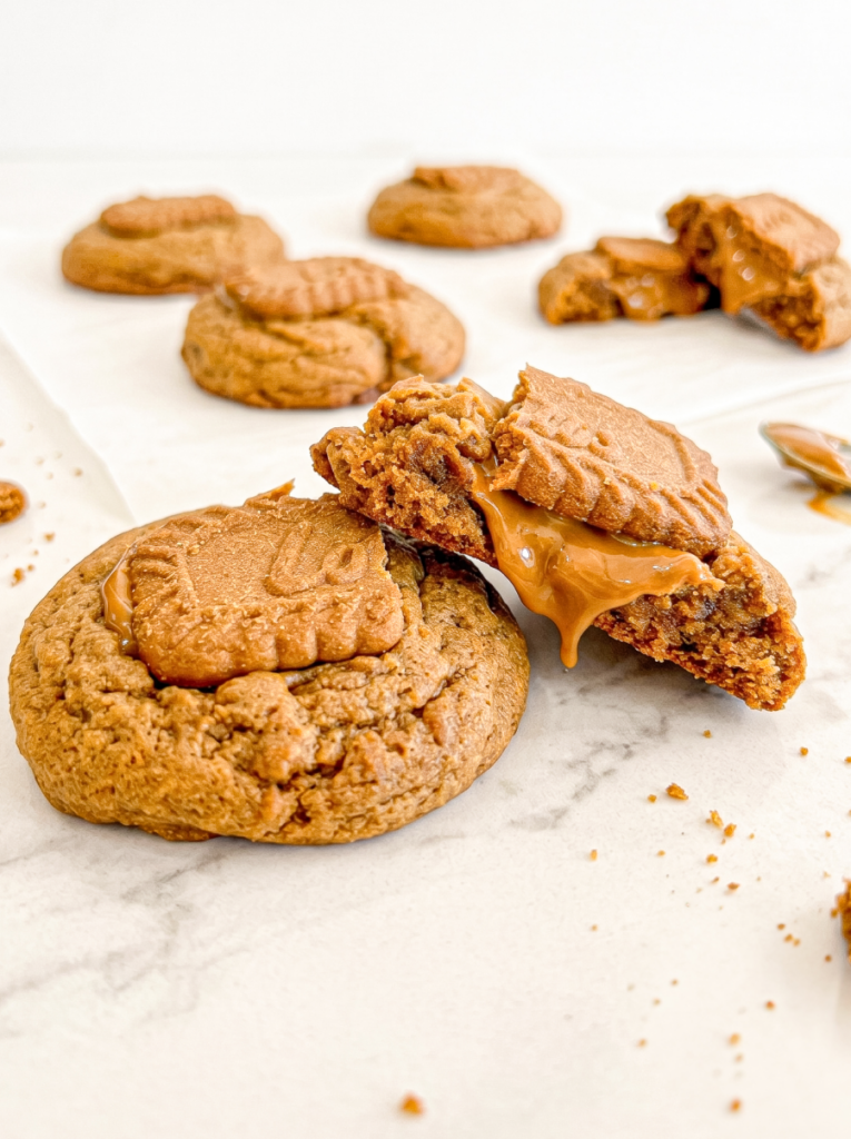 A broken gingerbread cookie with a Biscoff cookie on top shows a gooey Biscoff spread centre, with other cookies in the background.