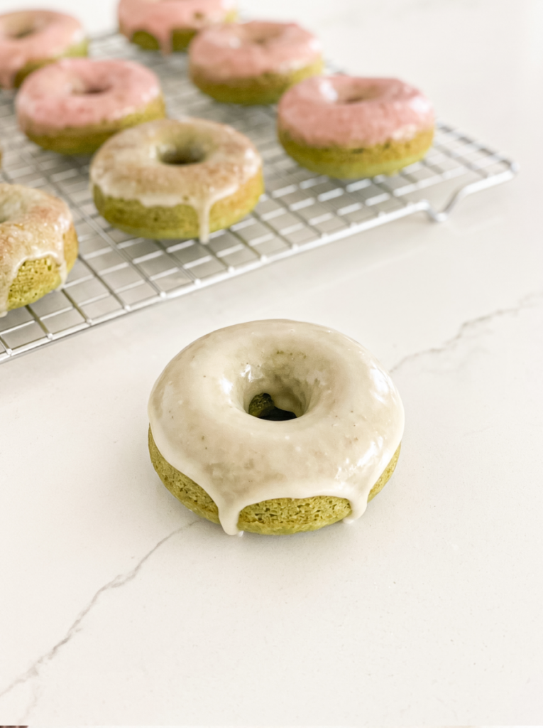 A green matcha donut with white glaze sits in the foreground, with more glazed matcha donuts on a wire rack behind it.