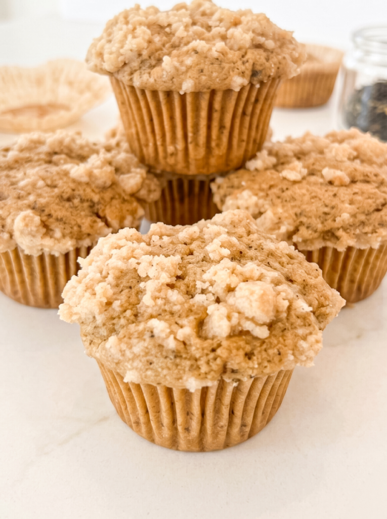 Four Earl Grey crumble muffins with streusel topping are stacked on a white surface, with a jar of loose tea in the background.