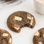 A brown hojicha latte cookie with white and milk chocolate chunks has a bite taken out, with other cookies and a latte.